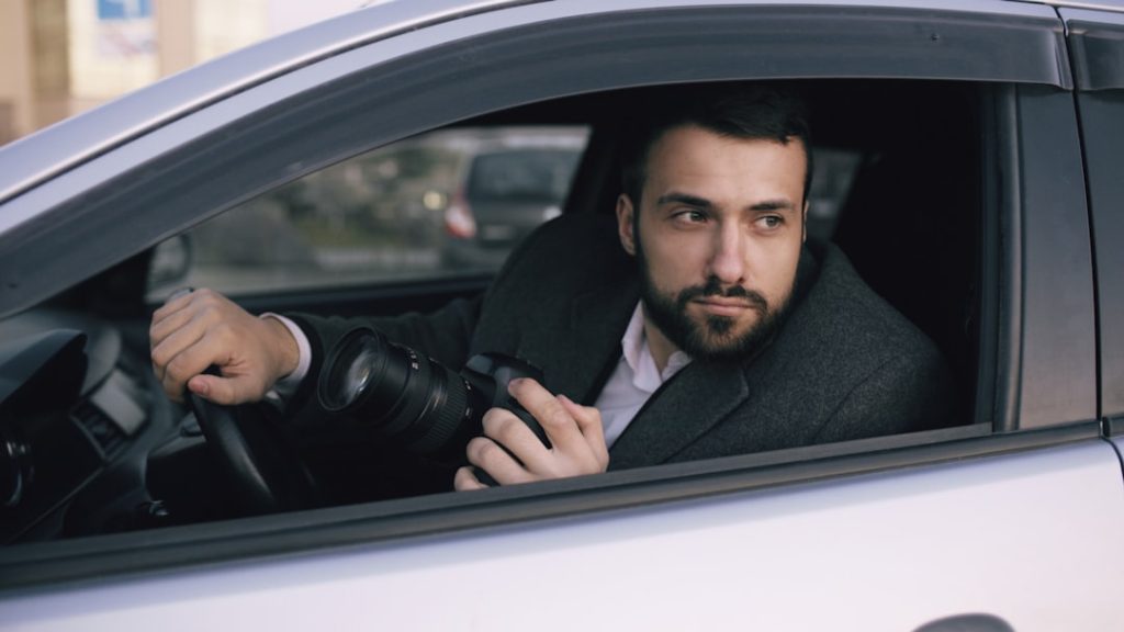 Young private detective man sitting inside car and photographing with slr camera