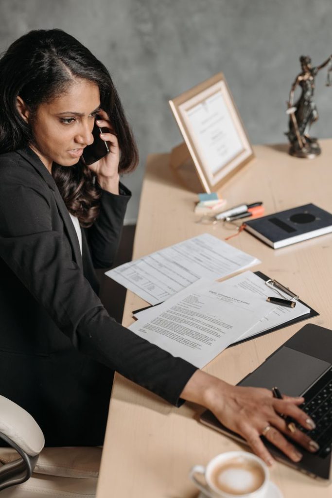 Professional businesswoman multitasking at an office desk, using a laptop and talking on the phone.